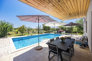 A swimming pool with a wooden pergola overhead, surrounded by a patio area with outdoor furniture and a view of greenery. - Villa Cvek (Kanfanar)