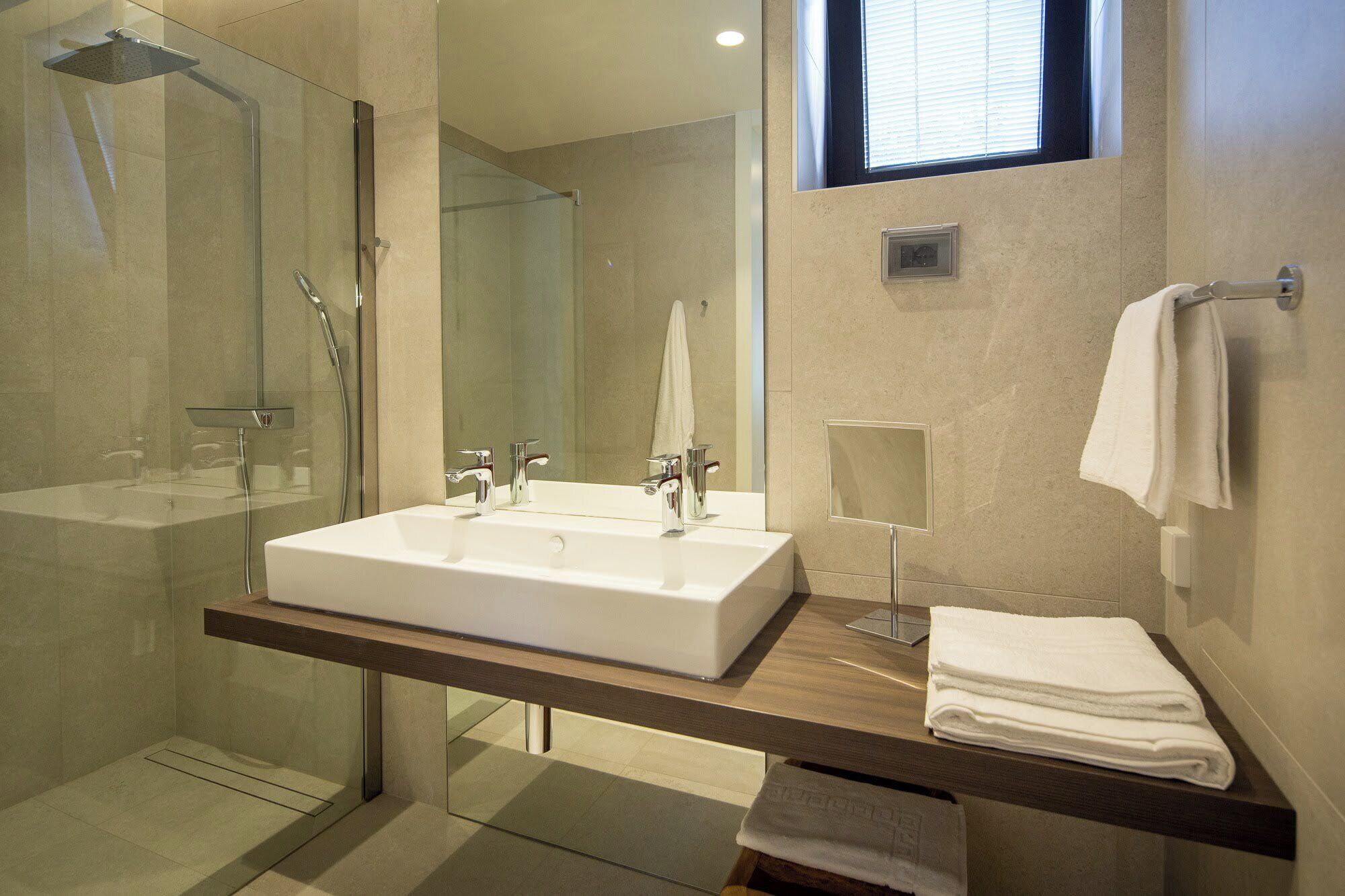 Een moderne badkamer met een douchecabine van glas, een groot wit wastafelmeubel en een houten paneel met een stap handdoeken.