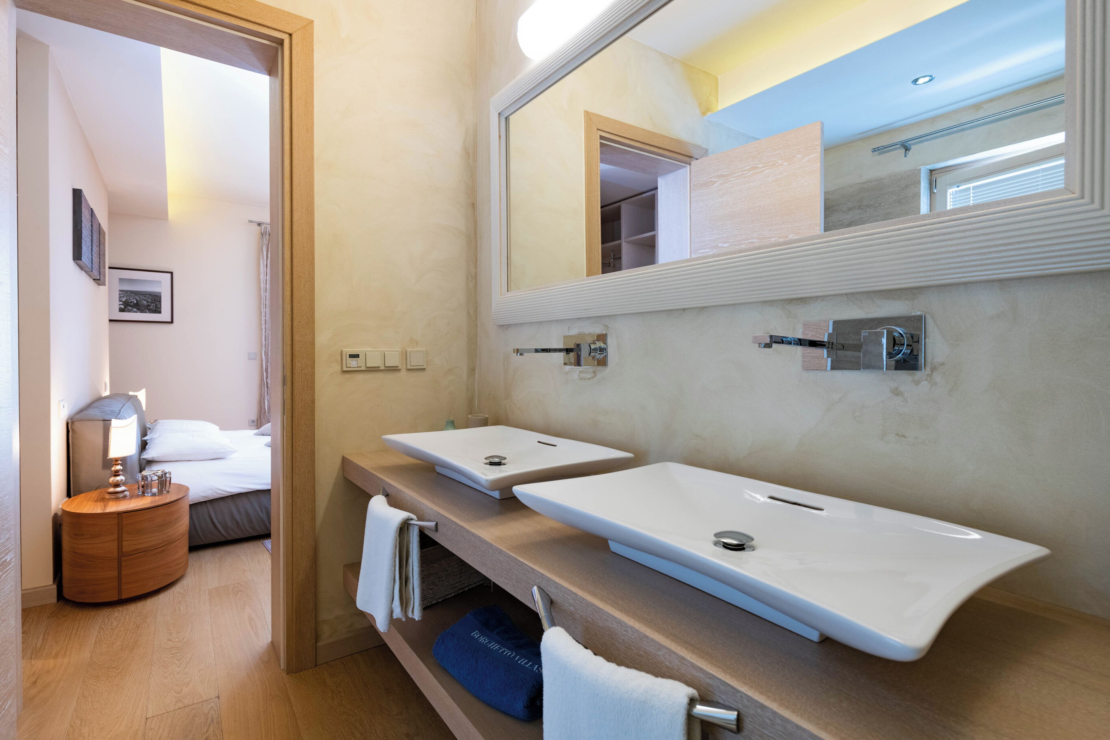 A modern bathroom with two sinks, a mirror, and a bedside table with a lamp.