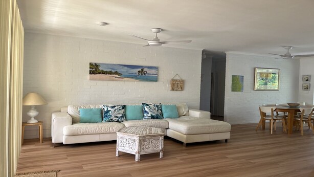 A living room with a white brick wall, a white sofa with blue and white cushions, a white coffee table, and a dining area with a wooden table and chairs. - Annette's - by Lifestyle. Brunswick Heads (BRUNSWICK HEADS)