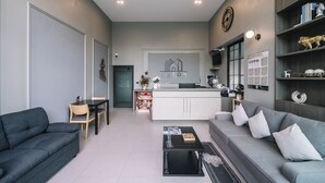 A modern living room with a grey sofa, a black coffee table, and a grey couch. There is a small dining area with a table and chairs. A kitchen area is visible in the background with a white countertop and a clock on the wall. - Aston Residence hatyai (Hat Yai)