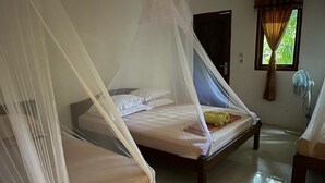 Une chambre avec deux lits, chacun doté d’un baldaquin. On y trouve une fenêtre avec des rideaux, une porte et un ventilateur fixé au mur.