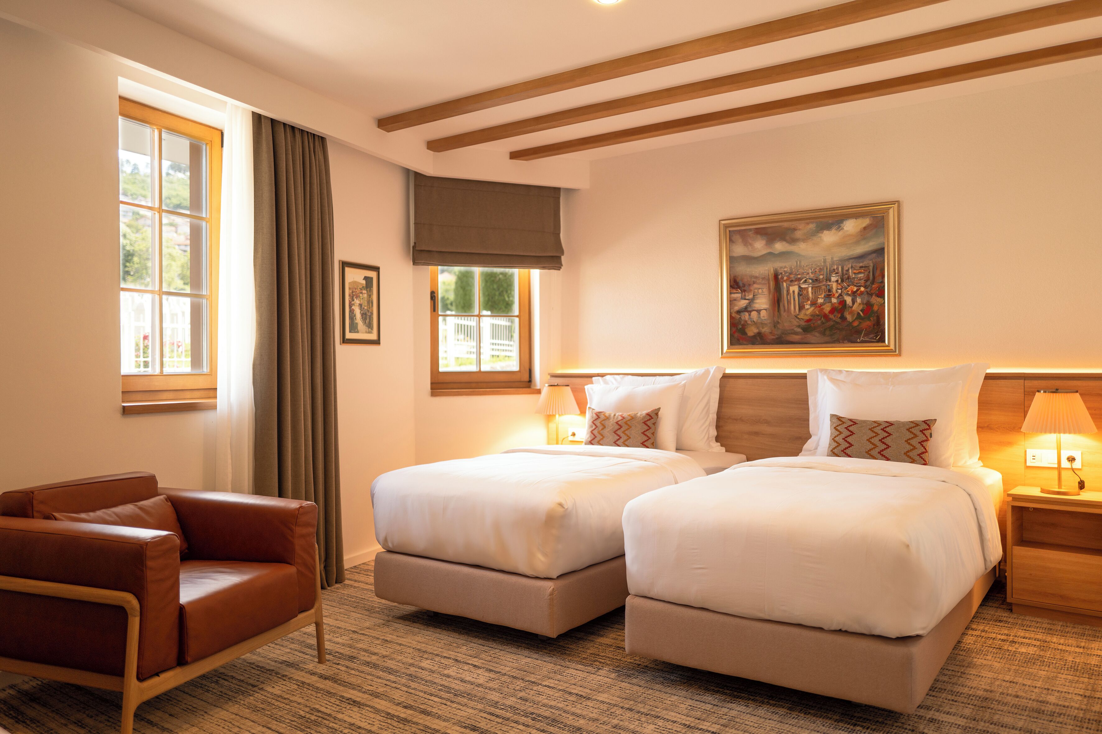 A hotel room with two beds, a brown leather chair, a painting on the wall, and a window with curtains.