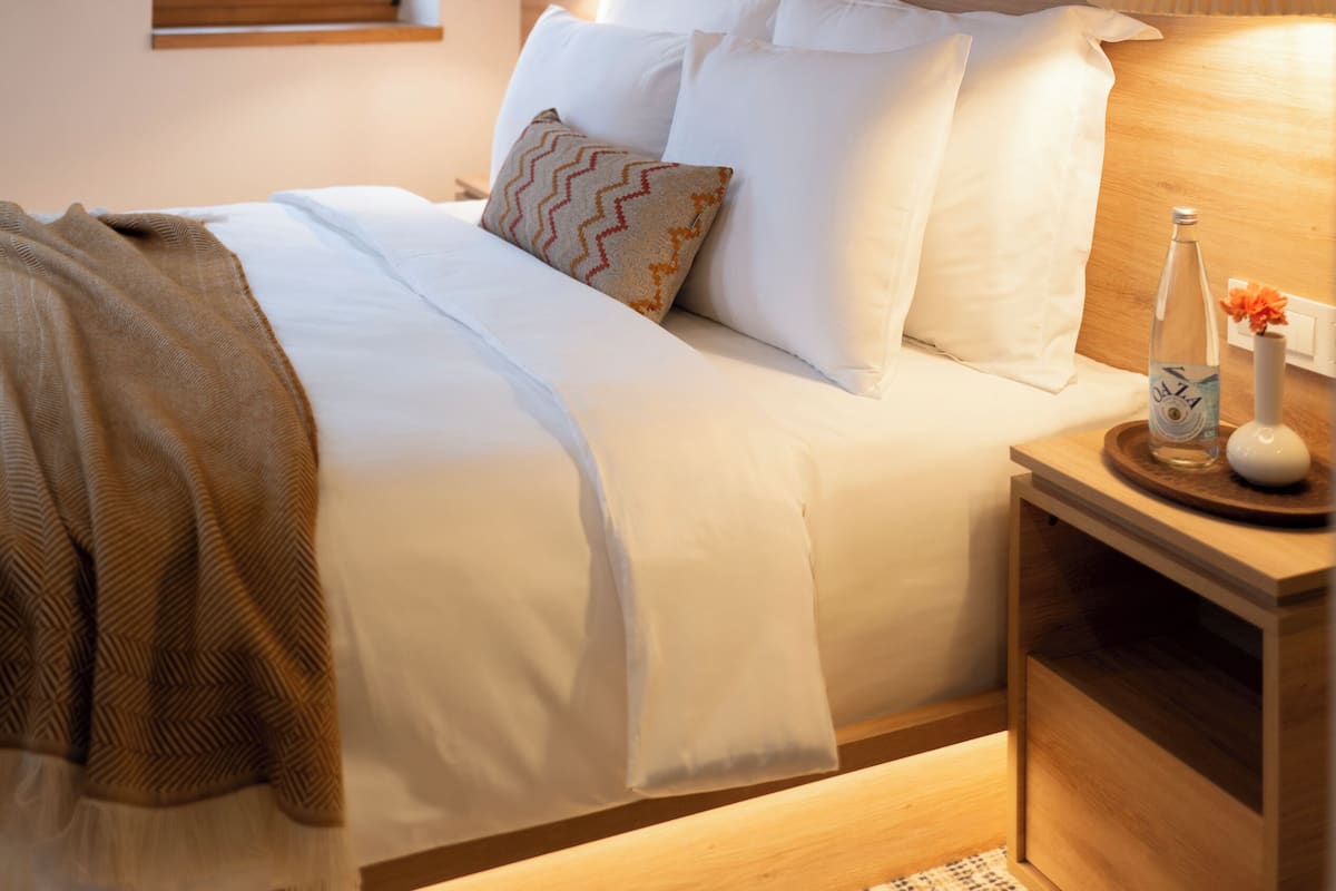 A neatly made bed with white linens and a patterned throw blanket, a bedside table with a bottle and vase, a painting above the bed, and a window with a view of greenery.