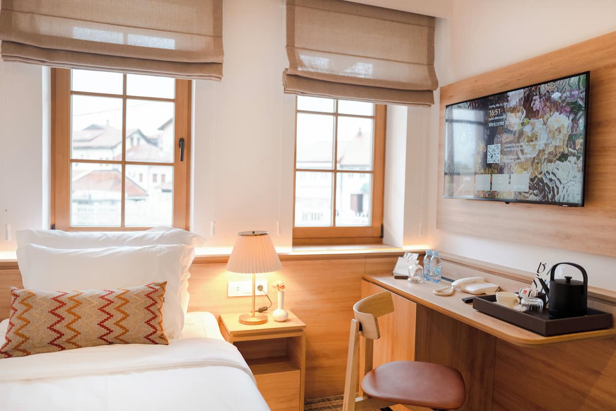 A hotel room with a bed, a desk, a chair, a lamp, a TV, and windows with blinds.