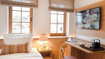 A hotel room with a bed, a desk, a chair, a lamp, a TV, and windows with blinds.