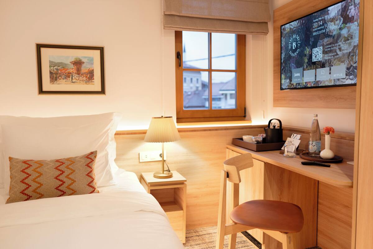 A hotel room with a bed, a desk, a chair, a lamp, a TV, and a window with a view of buildings.