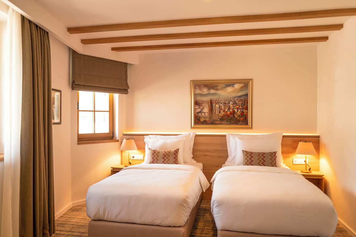 A hotel room with two beds, a painting on the wall, and a window with curtains.