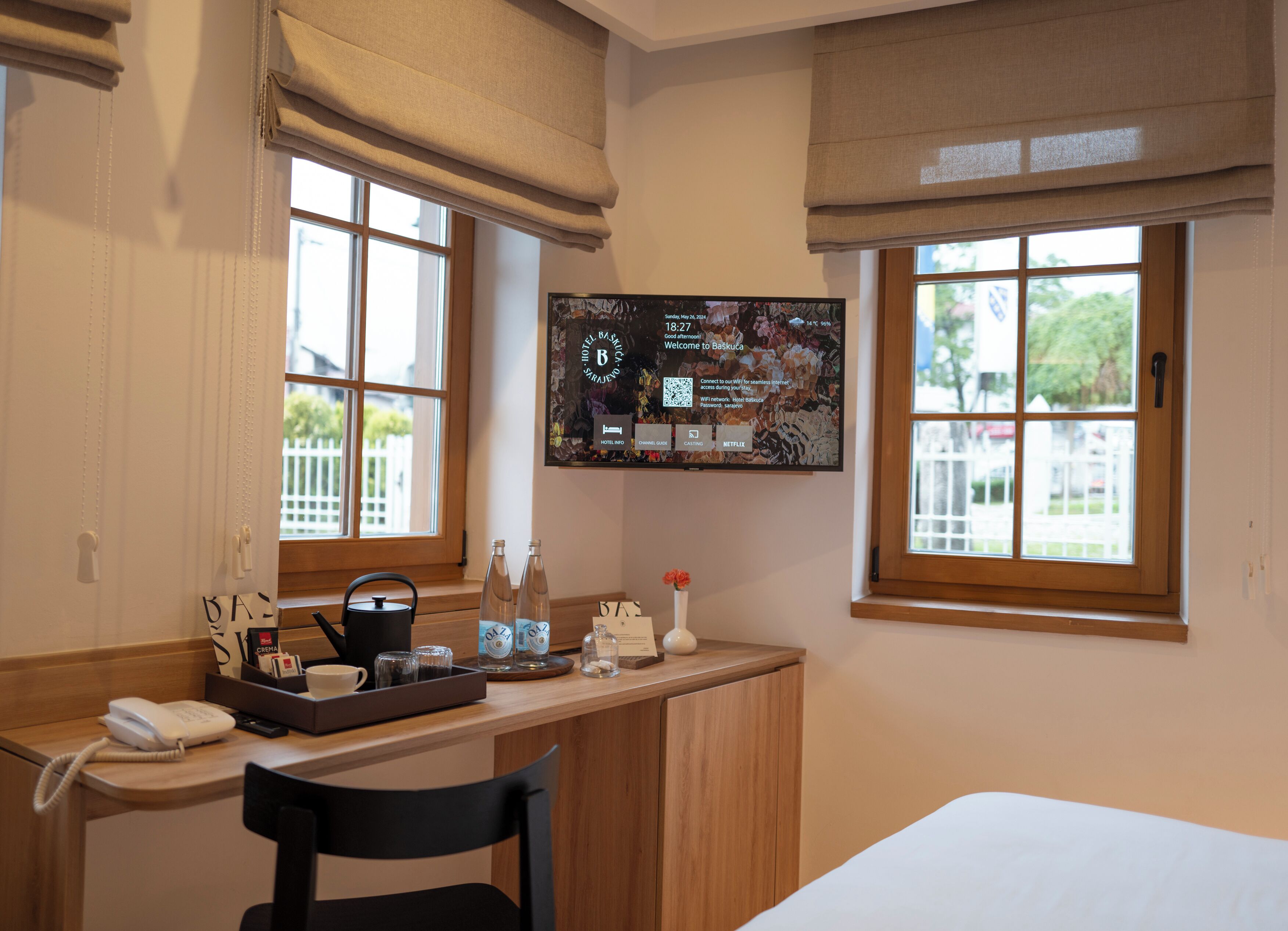 Habitación de hotel con escritorio de madera, televisor montado en la pared, cama y ventana con vista a una cerca blanca y follaje.