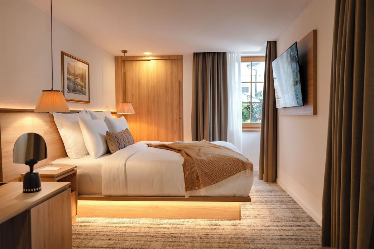 A modern hotel room with a large bed, bedside lamps, a flat-screen TV mounted on the wall, and a window with curtains.