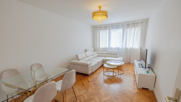 Una sala de estar moderna con una mesa de comedor de vidrio, sofás blancos, una araña y un televisor sobre un mueble blanco.