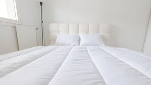 Una cama bien tendida con ropa blanca, un cabecero con diseño capitonado y una lámpara de pie a la izquierda.