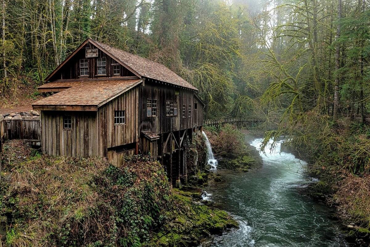 Una struttura in legno rustica con un tetto ripido, posata su palafitte sopra un fiume corrente, circondata da una fitta foresta.