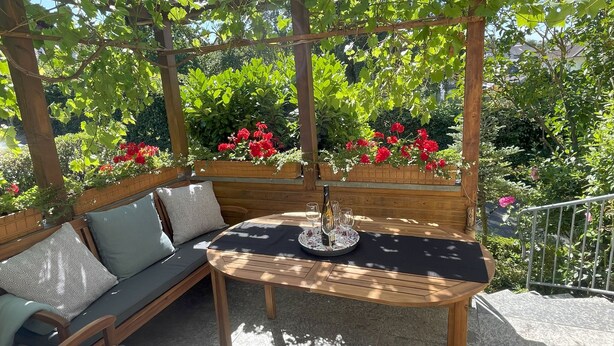 A wooden pergola with a table and a cushioned bench underneath. - 152 m² vacation apartment Quiet location on the edge of the village Overlooking the Rhine Valley Opposite the Loreley (Sankt Goar)