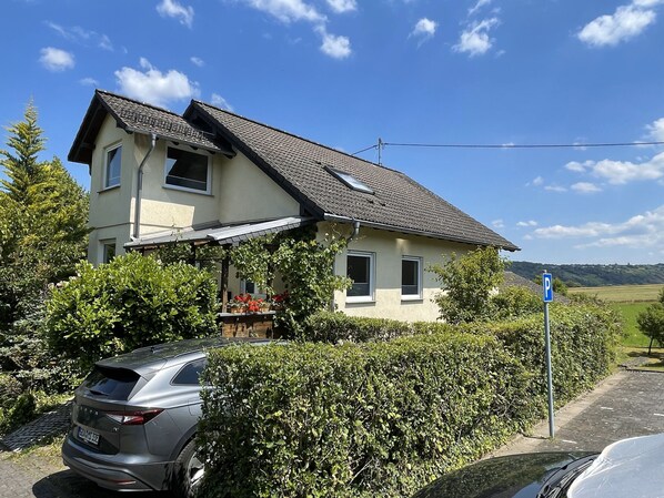 A two-story house with a sloped roof, a visible garage, and a parking area with a car. - 152 m² vacation apartment Quiet location on the edge of the village Overlooking the Rhine Valley Opposite the Loreley (Sankt Goar)