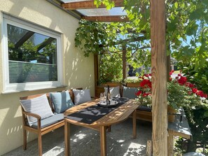 A wooden outdoor seating area with a table and chairs, a pergola with climbing plants, and a window with blinds. - 152 m² vacation apartment Quiet location on the edge of the village Overlooking the Rhine Valley Opposite the Loreley (Sankt Goar)