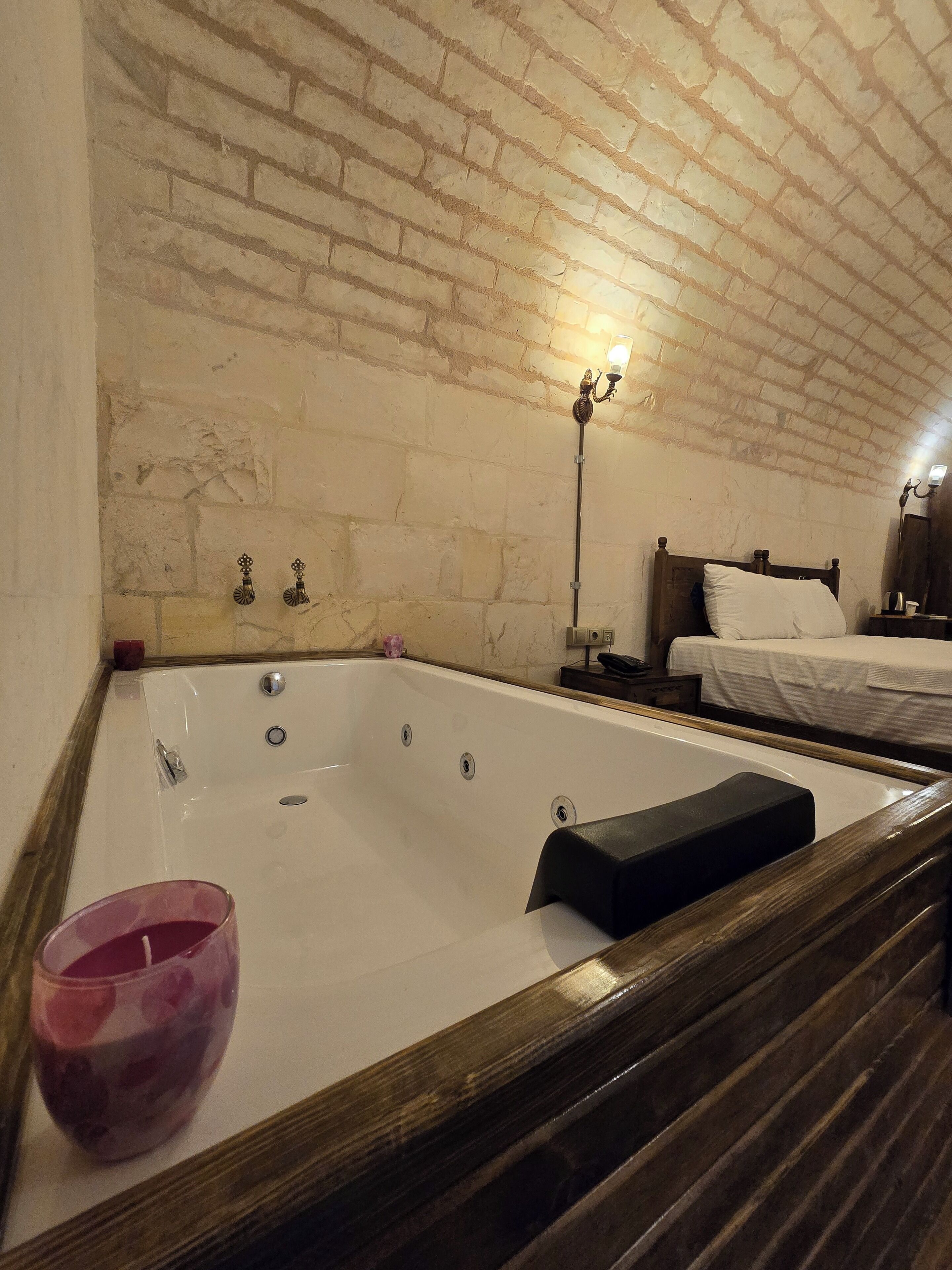 Una habitación con una bañera grande, un banco de madera, una cama con ropa blanca y una pared de ladrillo.