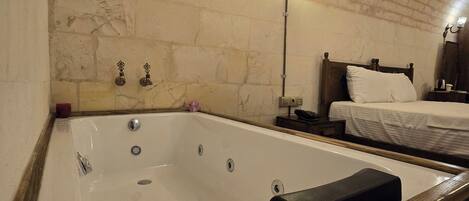 Una habitación con una bañera grande, un banco de madera, una cama con ropa blanca y una pared de ladrillo.