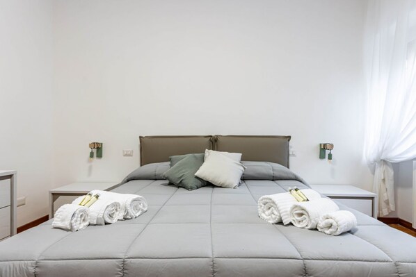 A neatly made bed with grey bedding, two pillows, and rolled white towels on either side.