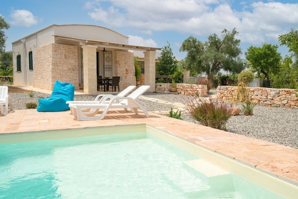 Un espace piscine avec des chaises longues, un bâtiment en pierre et un jardin agrémenté d’arbres et de plantes.