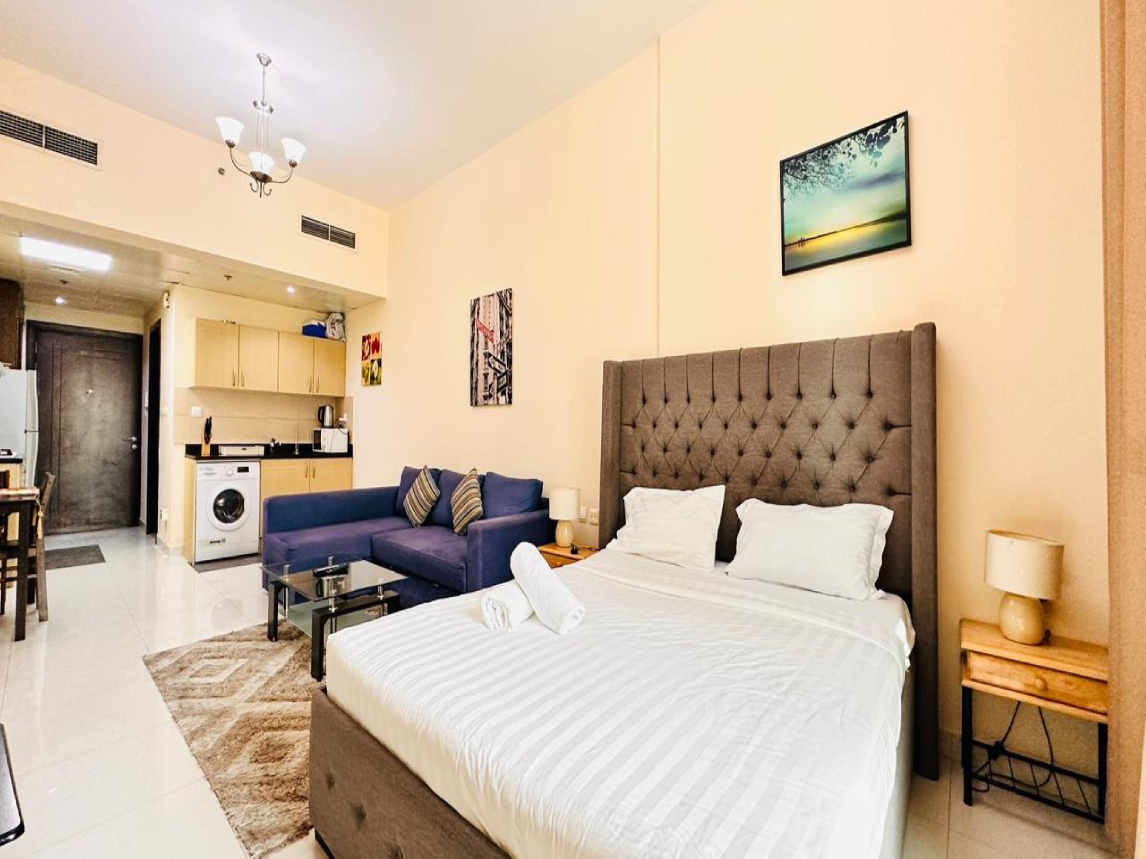 A modern hotel room with a large bed, a sofa, a small kitchen area, and a view of the cityscape.