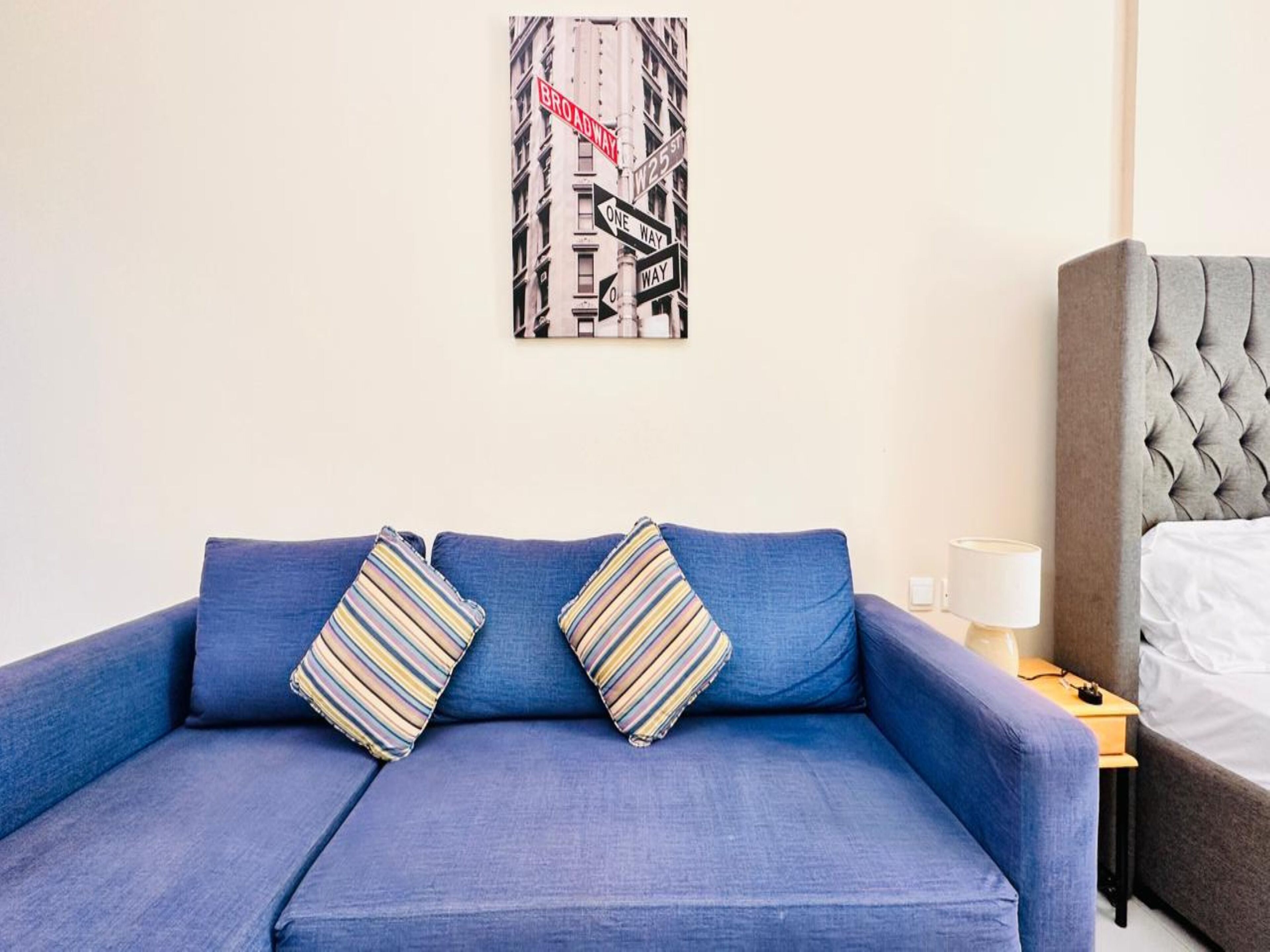 A room with a blue sofa, two striped pillows, a framed picture of a street sign, a bedside table with a lamp, and a grey headboard.