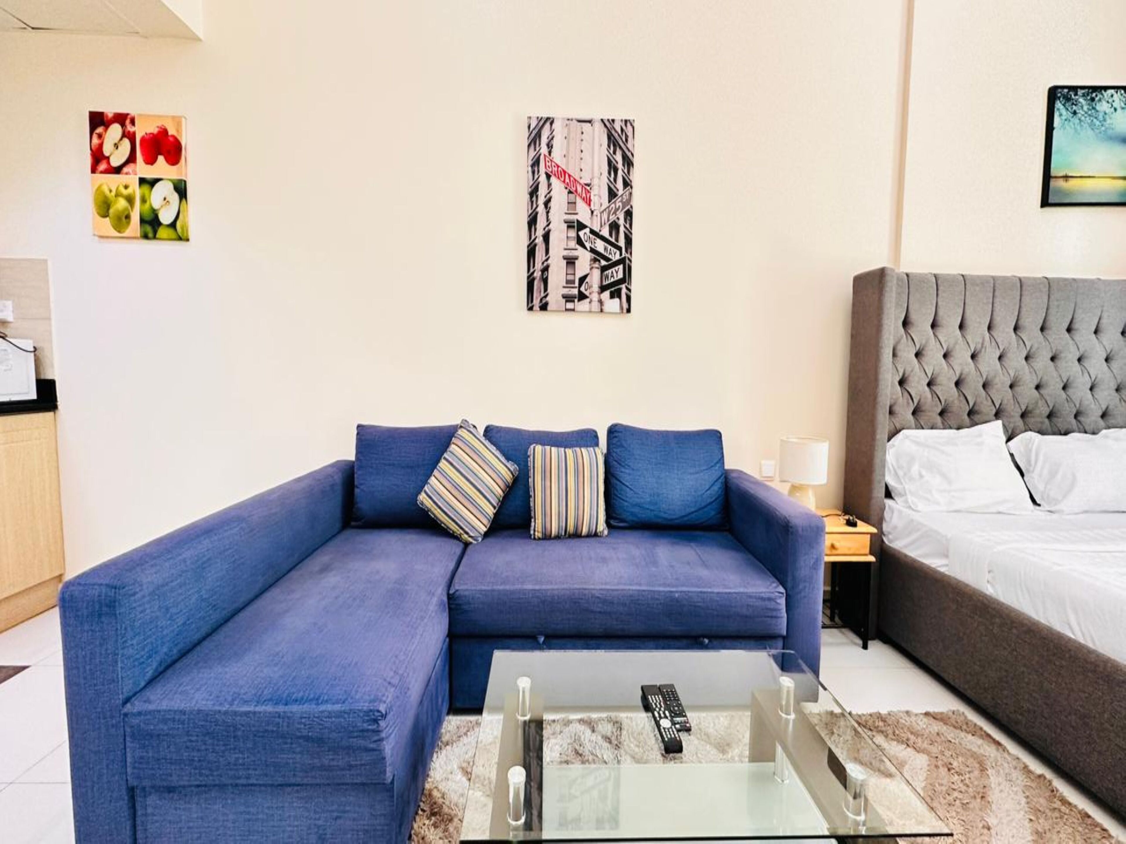 Un salon moderne avec un canapé bleu, une table basse en verre, un lit avec une tête de lit grise et une œuvre d’art accrochée au mur.