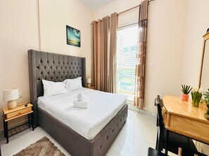 A hotel room with a large bed, a bedside table with a lamp, a small table with a plant, and a window with curtains.