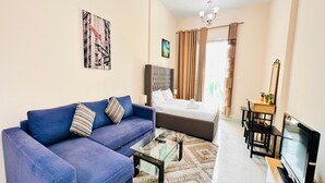 A modern living room with a blue sofa, a glass coffee table, a bed with a grey headboard, a TV, and a dining area with a table and chairs. - OYO 1233 Home Elite 1 Residence Sports City, Dubai (Dubai)