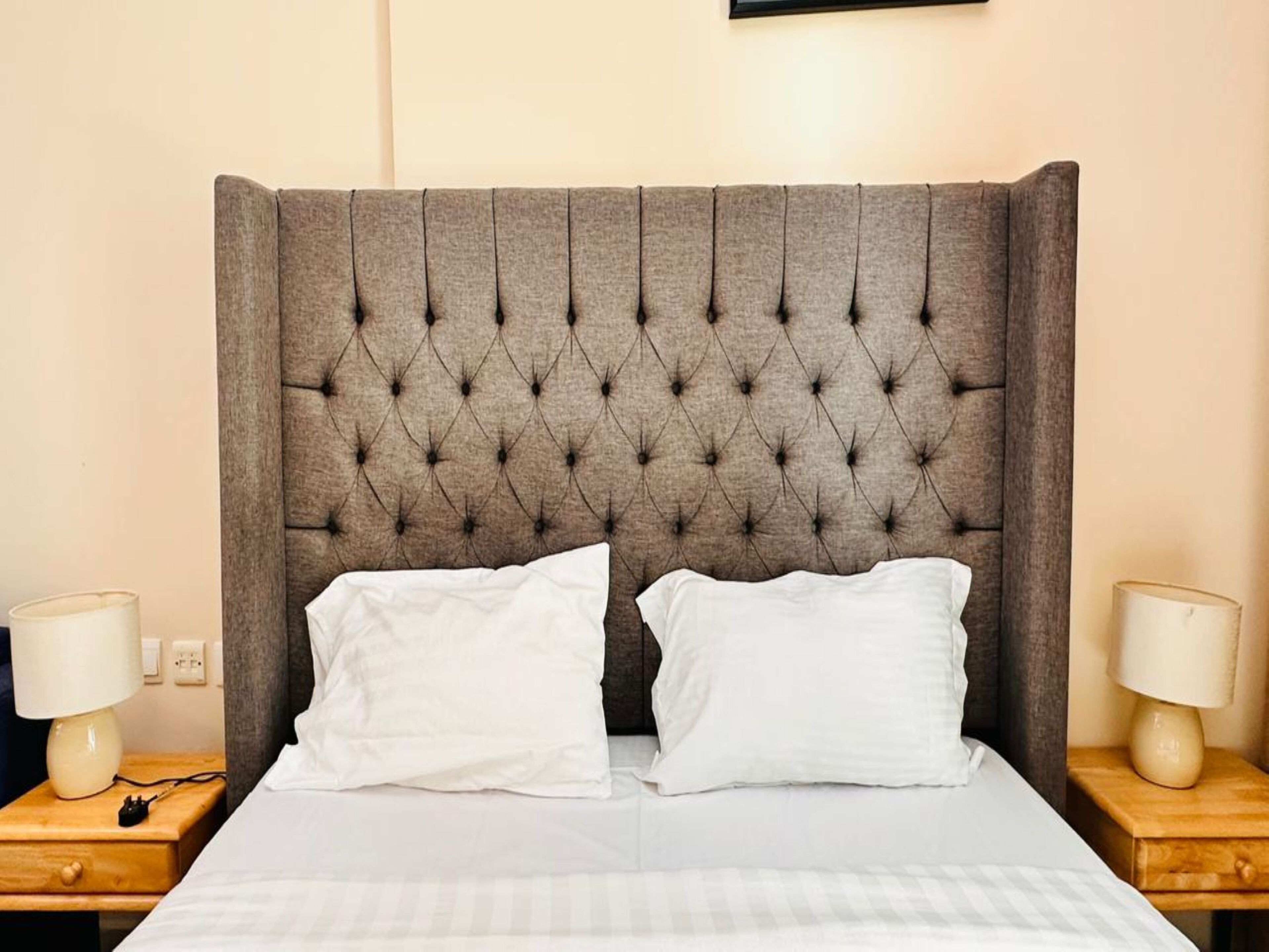 A bed with a tufted headboard, two white pillows, and a wooden nightstand with a lamp.