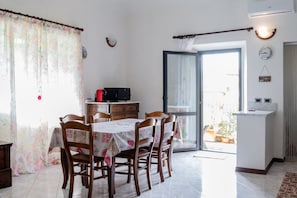 Een eetgedeelte met een tafel en stoelen, een keuken met een koelkast, en een schuifpui die leidt naar een buitenruimte.