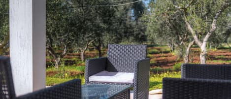 An outdoor seating area with wicker furniture and a glass table, surrounded by greenery and trees.