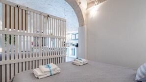 A hotel room with a white brick wall, a wooden slat partition, a bed with a quilted grey bedspread, and a neatly made bed with towels. - Il Vico 8 Ostuni by Rentbeat (Ostuni)