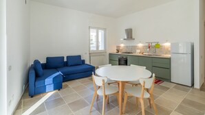 Ett kompakt bostadsutrymme med ett litet kök, en blå soffa, ett runt matbord med stolar och ett fönster som släpper in naturligt ljus.