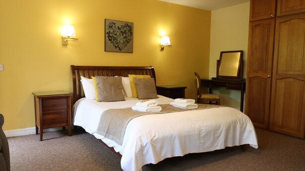 A neatly made bed with pillows and a blanket, a bedside table with a lamp, a wooden wardrobe, and a framed picture on the wall. - St George's Lodge (Woking)