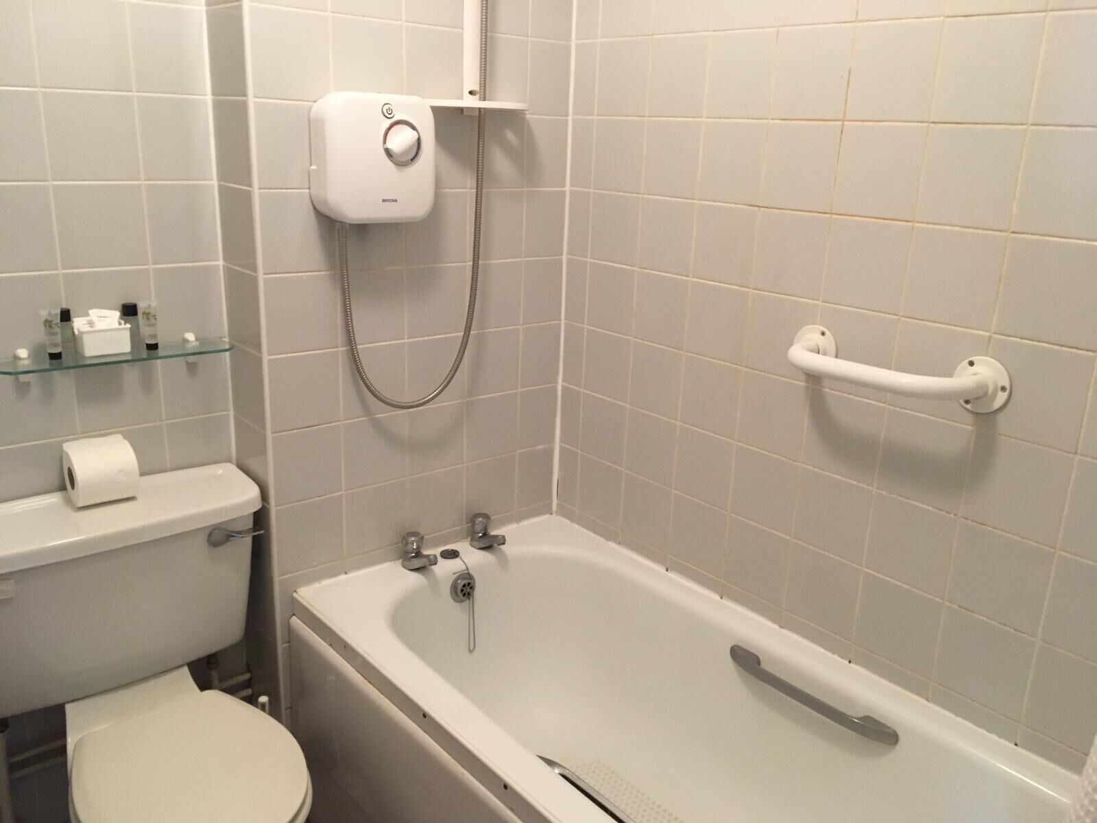 A bathroom with a toilet, a glass shelf with toiletries, a shower with a handheld showerhead, and a bathtub with a grab bar.