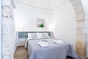 A bedroom with a bed, pillows, a nightstand, a small shelf with books and a bottle, and a stone wall. - Il Vico Suite Ostuni by Rentbeat (Ostuni)
