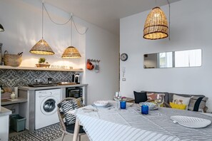 Kompakti keittiö, jossa on pesukone, ruokailutila kahdelle katetulla pöydällä sekä olohuone, jossa on sohva ja suuri peili.