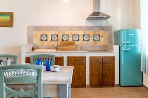 Une cuisine avec un mur recouvert de carreaux, des meubles en bois, un réfrigérateur de couleur turquoise et un coin repas avec des chaises et une table.