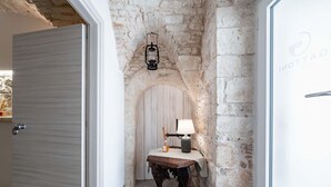 Un couloir aux murs de pierre, avec une porte en bois, une petite table sur laquelle se trouve une lampe, et une lanterne accrochée au mur.