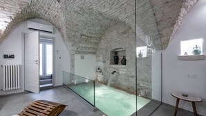 Une pièce aux murs de pierre, dotée d’une piscine entourée de parois en verre, d’un banc en bois et d’une petite table.