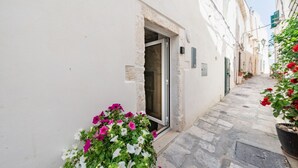 A narrow street with a building entrance, a potted plant with flowers, and a clear sky. - Donna Alfa Suite by Rentbeat (Ostuni)