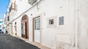 Eine enge Straße mit Gebäuden, ein geschlossener Laden mit einer braunen Tür sowie eine weiße Wand mit kleinem Fenster und Verteilerkasten.