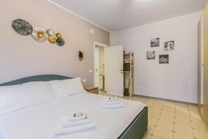 A neatly made bed with folded towels, a wall clock, and a door leading to a bathroom.