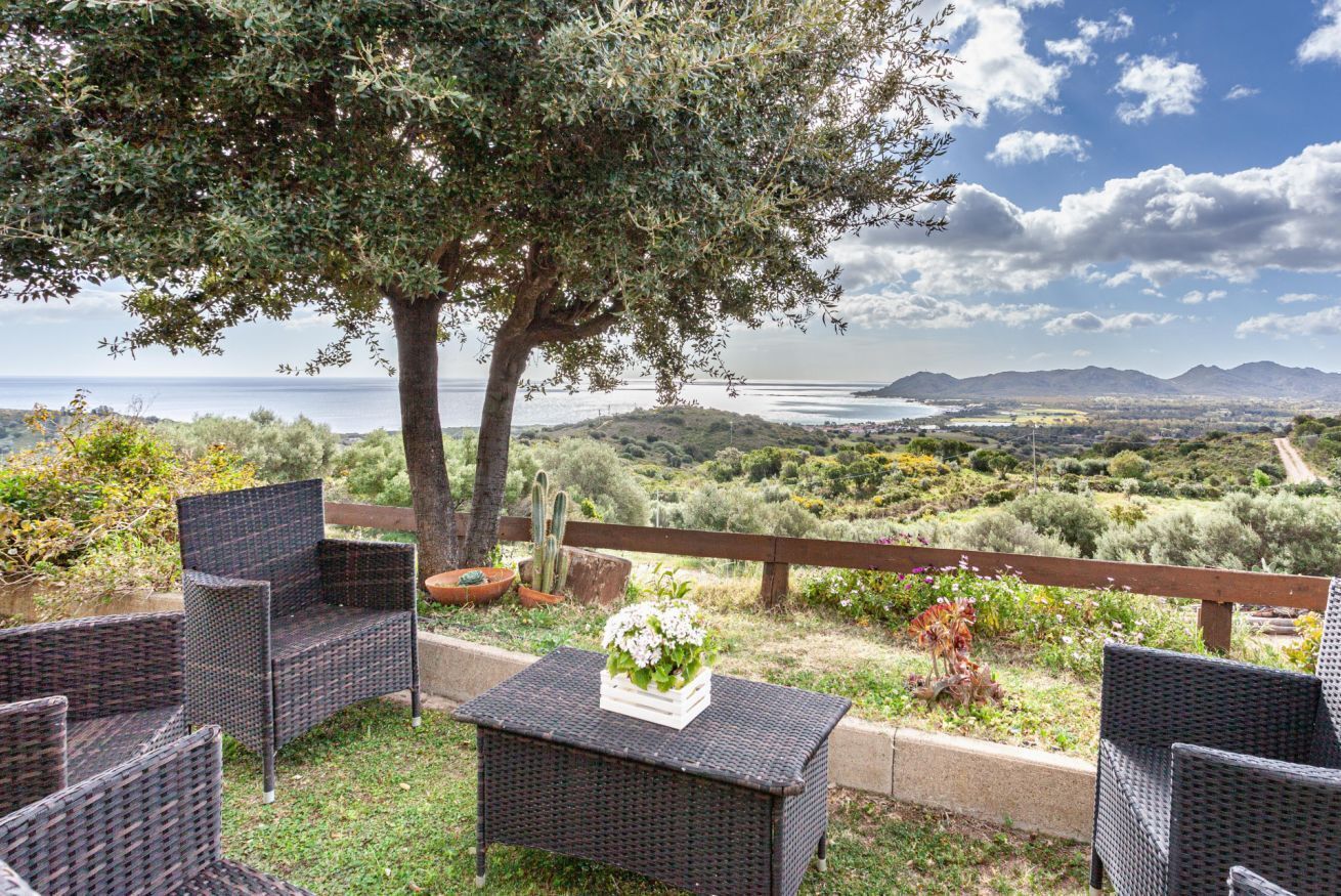 Ein Außenbereich mit Korbmöbeln, einem Tisch mit Blumen und Blick auf das Meer und die Berge.