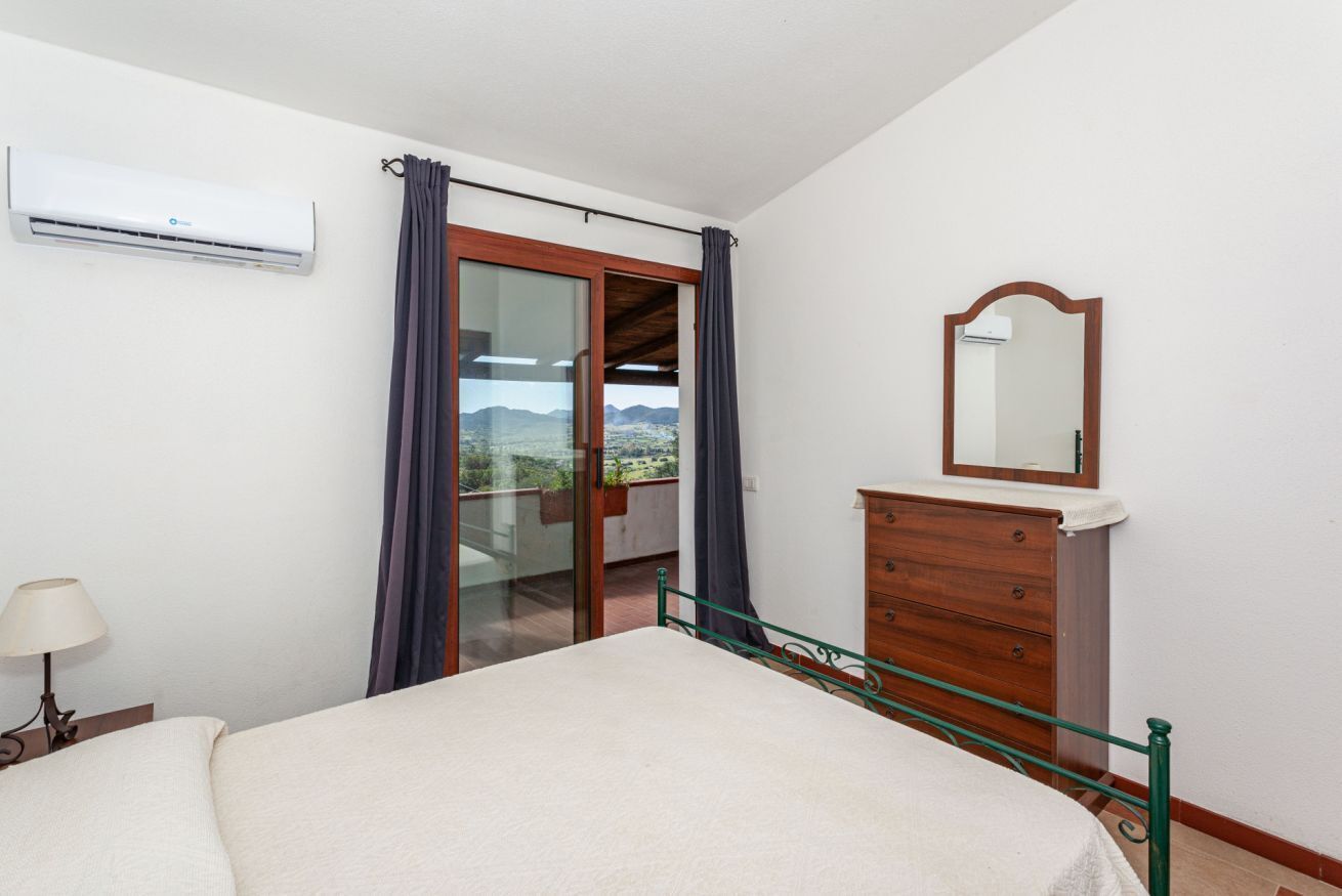A bedroom with a bed, a dresser, a mirror, and a sliding glass door leading to a balcony with a view.