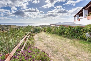 Un sentier de jardin mène à un bâtiment blanc surmonté d’un toit rouge, entouré de verdure et offrant une vue sur la mer.