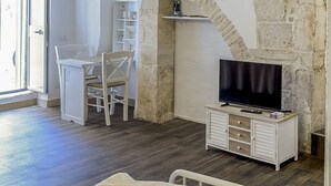 A room with a stone archway, a television on a wooden cabinet, a white chair, and a white shelf unit.