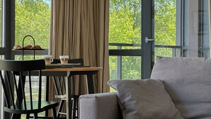 Ein modernes Zimmer mit einem grauen Sofa, einem Holztisch mit einer Tasse und Gebäck, und Blick auf Bäume durch große Fenster.