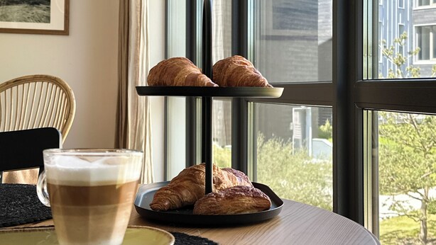 Ein Tisch mit einer Auswahl an Croissants und einer Kaffeetasse, ein Korbstuhl und Blick auf Gebäude durch große Fenster.